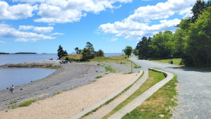 Wide gravel coastal walking trail, Sailor's Memorial Way, at Point Pleasant Park in Halifax, Nova Scotia, overlooking the sandy and rocky shoreline of Black Rock Beach with the Atlantic Ocean under a bright blue sky.