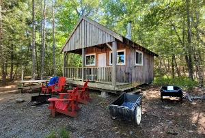 A rustic wooden cabin (C2) at Jeremy's Bay campground in Kejimkujik National Park, Nova Scotia, featuring a front porch, red Adirondack chairs, a fire pit, and a picnic table nestled in a lush green forest.