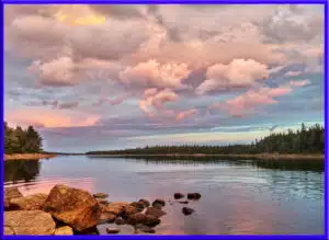 Panoramic sunset over Long Lake in Halifax, Nova Scotia, featuring calm reflective water, a rocky shoreline in the foreground, and large pink-tinted cumulus clouds filling the sky over a distant evergreen forest in Long Lake Provincial Park.