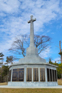 The Halifax Memorial at Point Pleasant Park, a large stone cross monument with bronze name plaques honoring sailors, soldiers, and merchant marines lost at sea, set against a bright blue sky in Halifax, Nova Scotia.