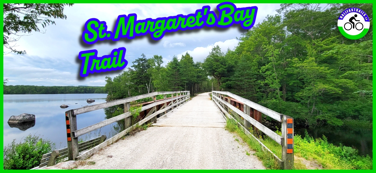 A scenic, wide-angle view of the St. Margaret's Bay Trail on a bright, partly cloudy day. A flat, gravel multi-use trail leads onto a wooden bridge with rustic timber railings spanning a calm, blue lake. The left side of the bridge overlooks the peaceful water with scattered boulders, while the right side is bordered by a lush, vibrant green forest. Stylized text in the top left corner reads "St. Margaret's Bay Trail" in a bright green font with a blue outline. A small, circular logo featuring a bicycle icon and the website "halifaxtrails.ca" is centered at the bottom.