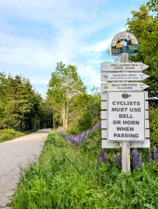 St. Margaret's Bay Rails-to-Trails gravel path lined with purple lupines, featuring a trail signpost with directions to local businesses and a cyclist bell warning sign.