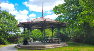 An ornate Victorian-style green metal gazebo with a red-trimmed roof and picnic tables inside, situated along a paved walking path surrounded by lush green trees at Point Pleasant Park in Halifax, Nova Scotia.