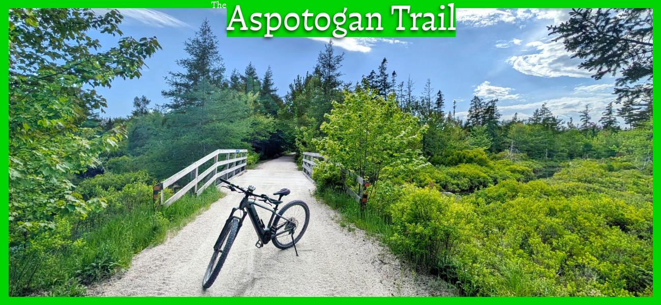 A bicycle parked on a wooden bridge along the Aspotogan Trail in Chester, Nova Scotia, with lush green foliage and blue sky, featuring a title graphic